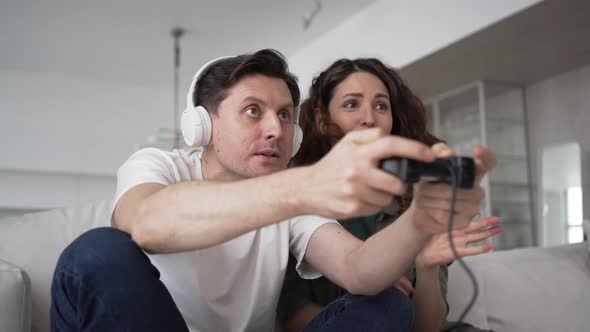 A Man Plays a Video Game with Headphones and a Gamepad in His Hands His Wife Sits Next to Her and