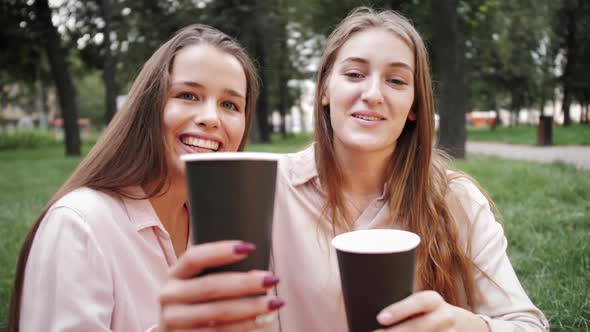 Young stylish cheerful girls having picnic at nature while sitting on grass and drinking beverages.