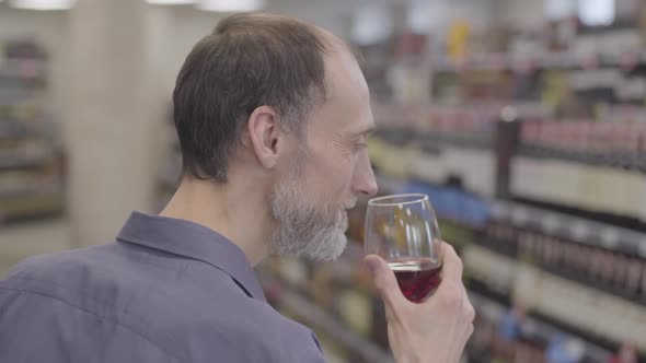 Side View Close-up of Adult Caucasian Sommelier Shaking and Smelling Red Wine in Glass. Smiling