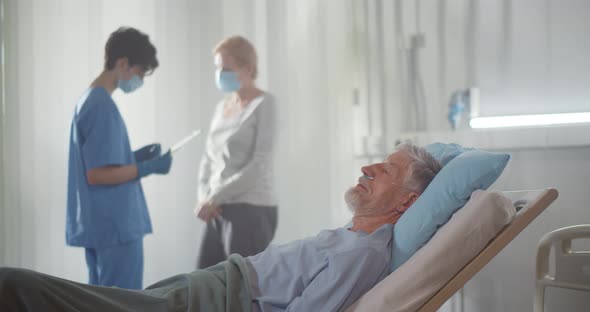 Sick Aged Man Lying in Hospital Bed While Wife and Doctor Discussing Diagnosis on Background