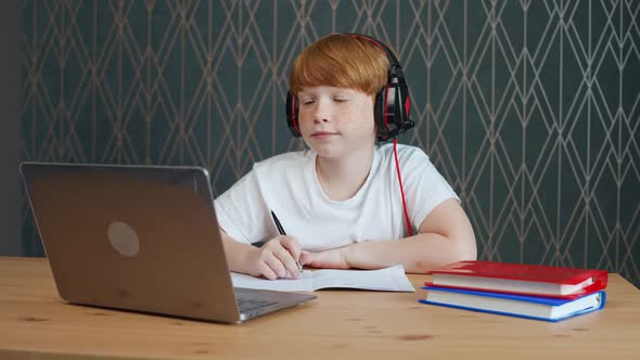 Distance Learning Redhaired Teen Boy Sitting on the Table in Living Room and Uses a Laptop to Study
