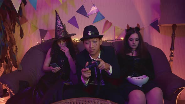 The Guy in the Creepy Costume is Actively Playing the Console and the Girls are Sitting on the Phone