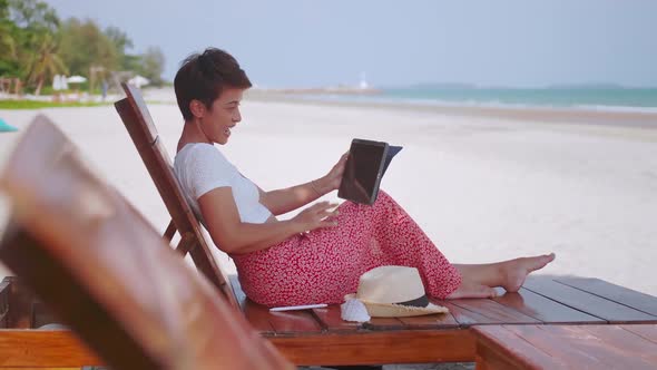 Asian woman chatting with family on video call while relaxing on beach.