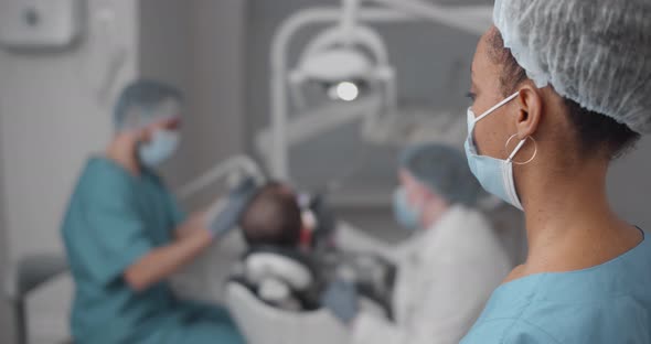 African Woman Intern Standing in Dental Office Watching Professional Dentist Treating Patient Teeth