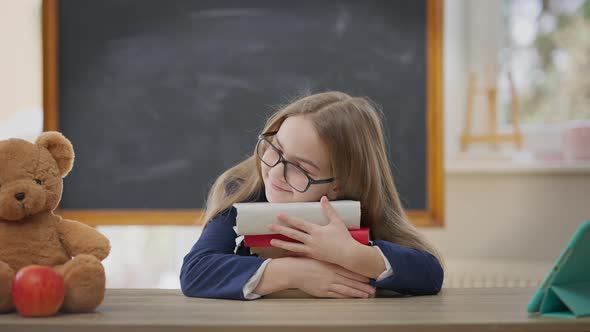 Smiling Schoolgirl in Eyeglasses Hugging Books Sitting at Desk in Classroom and Looking at Camera