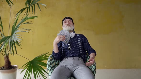 Senior Bearded Man Sitting in Chair Near Yellow Wall with Palm Tree and Waving with Dollars 