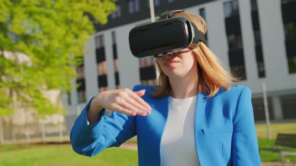 A Happy Woman in the Suit is Standing Outside and Gestures in Augmented Reality in 3D VR Goggles