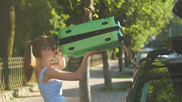 Young Female Tourist Taking Out Green Suitcase From Car Roof Rack and Walking on City Street