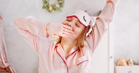 Young yawns woman in pink pajamas and sleep mask, waking up and stretching after sleep