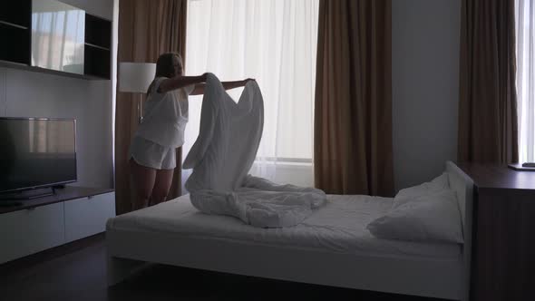 Happy Blonde Woman Makes the Bed in the Bedroom Spreads White Blanket on the Bed