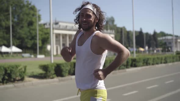 Portrait of Happy Caucasian Sportsman Walking Fast on Sunny City Street, Smiling at Camera, and