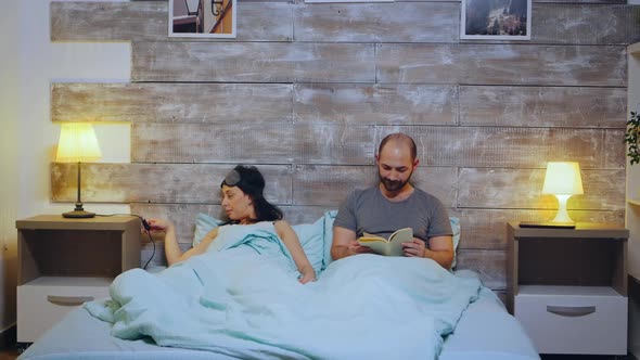 Young Man Reading a Book While His Wife Is Turning the Lights Off