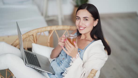 Woman Sits in Chair with Laptop, Holds Cup of Tea in Her Hands, Drinks It, Smiles