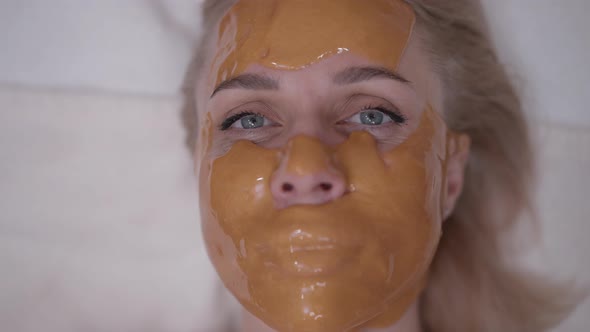 Closeup Slim Blond Caucasian Woman in Yellow Facial Mask Looking at Camera with Grey Eyes Smiling