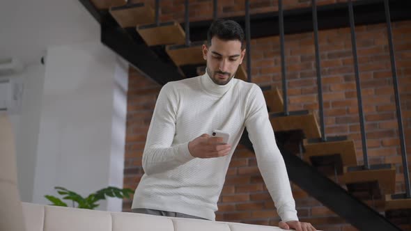 Thoughtful Confident Young Man Messaging on Smartphone Standing in Home Office Indoors