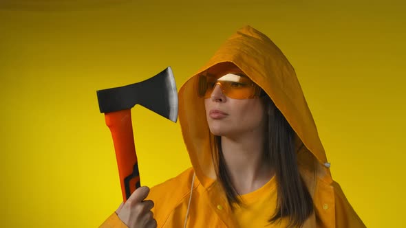 A Young Beautiful Woman in a Yellow Raincoat Examines an Axe in Her Hand