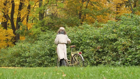 A Cute Young Woman in a Beige Raincoat Walks with a Bicycle Through the City Autumn Park Back View