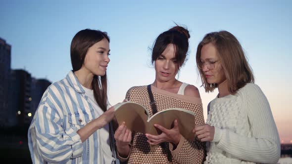 Women Standing Outdoors Looking at Magazine Discussing It.