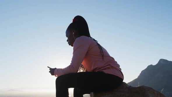 African american woman exercising outdoors sitting on rock using smartphone in countryside at sunset