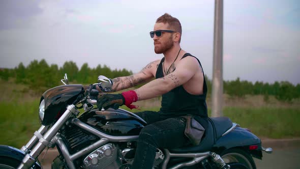 Stylish Biker with Tattoos Rides a Motorcycle on a Country Road at Sunset