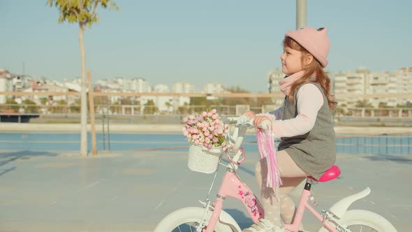 Little Cute Girl On A Pink Bicycle With Flowers Rides In The Park