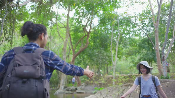 Young couple Asian man help Caucasian woman friend crossing river traveling in the forest together.