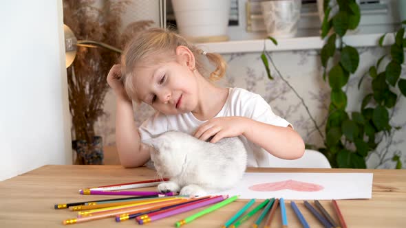 a Little Girl is Stroking a White Kitten That is Playing with Colored Pencils on a Wooden Table