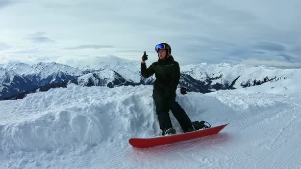 Teenage snowboarder taking selfie sitting on a snow surrounded by mountains.