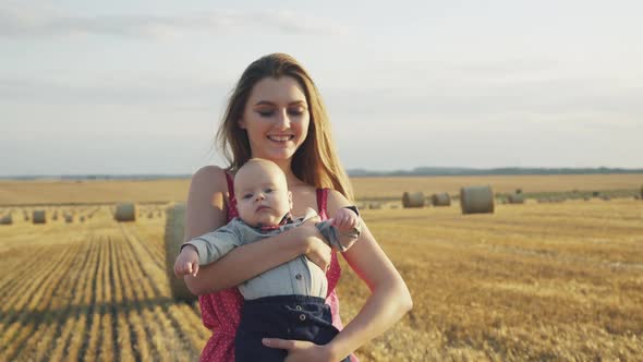 Portrait of Happy Mother Lulls Her Lovely Baby in Hands with Smile in Hay Field