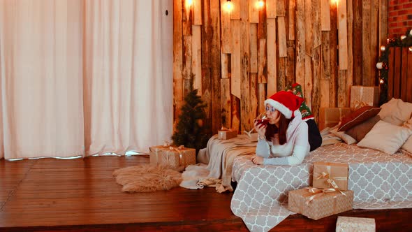 Young Woman in Santa Hat and Glasses Lying on Bed and Drinking Red Wine