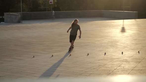 Young Long-haired Man Roller Skater Is Dancing Between Cones in the Evening in a City Park at Sunset