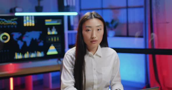 Asian Woman which Working on Computewr in Evening Office and then Looking Into Camera