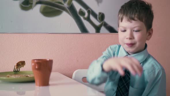 CU Schoolboy Sits at the Table in School Uniform and Has Breakfast Playing Before Going to School