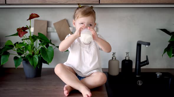 Happy Baby Girl Sitting at the Table in the Kitchen and Drinking Milk