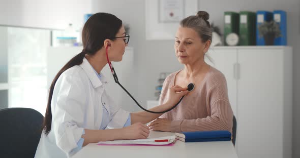 Young Female Doctor in White Coat Listening with Stethoscope to Aged Patient in Office