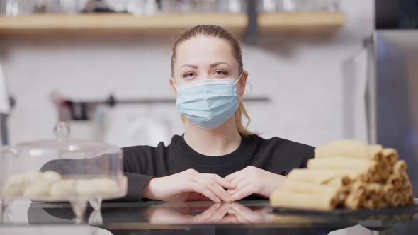Portrait of Positive Smiling Young Woman in Covid Face Mask Standing at Counter with Sweet Desserts