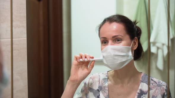 Portrait of Caucasian girl in night robe wearing face mask, brush teeth, look in mirror in bathroom.