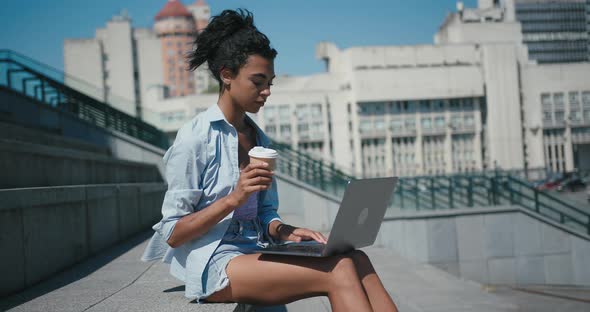 Black Woman with Cup Types on Laptop Sitting on Stone Steps