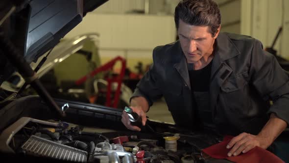 Auto mechanic standing at the opened hood and lighting with lamp while working at the workshop