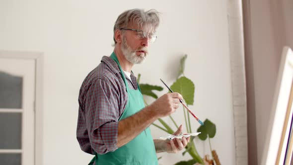 Male Artist in an Apron Paints on Canvas Dips Brush Into Palette Works in Workshop Front View