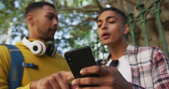 Two happy mixed race male friends standing, talking and using smartphone in the street