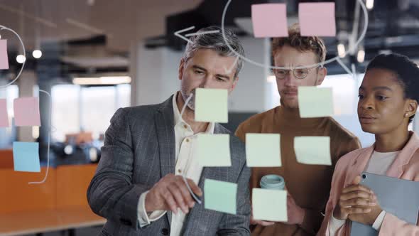 Senior Male Office Worker Using Glass Board and Proposing His Problem Solving to Workmates