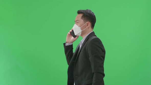 Asian Business Man Talking On Mobile Phone While Walking On Green Screen, Wear Medical Mask