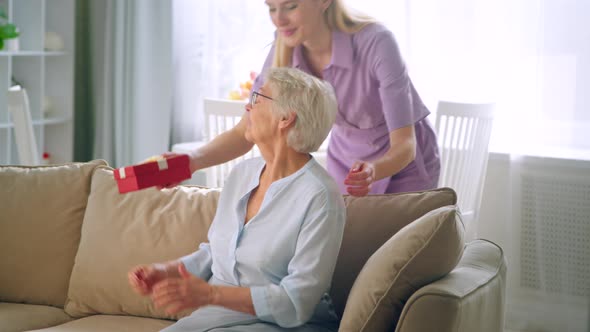 Long haired blonde gives small box with present to aged lady with joyful smile at birthday