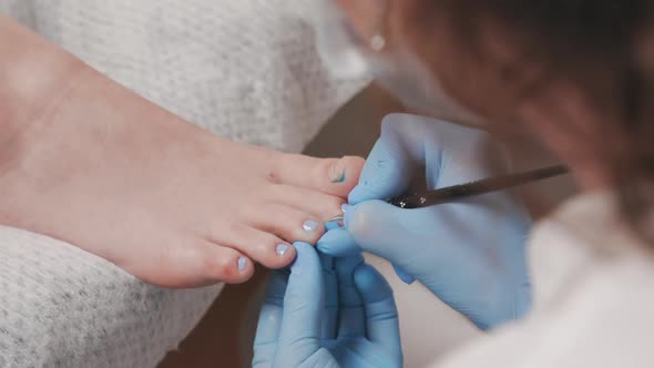 Pedicurist in medical gloves paints the client's toenails with blue nail polish. Closeup