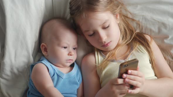 Two Children Use and Play Mobile Phone Together on Their Bed. Kids Use Telephone Together on Bed in