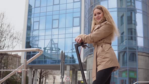 Young Stylish Blonde Girl in a Beige Trench Coat and Jeans Stands with an Electric Scooter on the