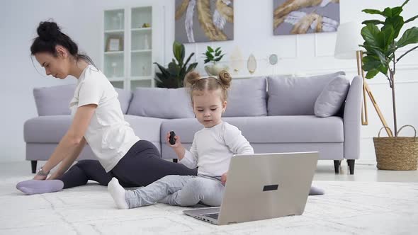 Small Girl Sitting Near the Laptop on the Floor while Her Confident Sportive Dark Haired Mother 