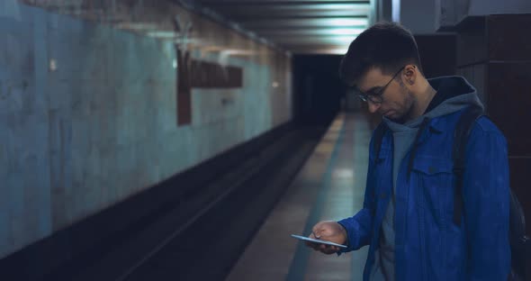 Young Man in Glasses is Standing at the Metro Station and is Looking at the Phone