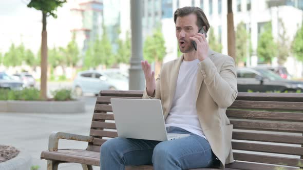 Angry Young Man with Laptop Talking on Phone While Sitting on Bench